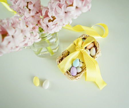 Treat candy for Easter in basket with an yellow bow and glass vase with hyacinths on white table....