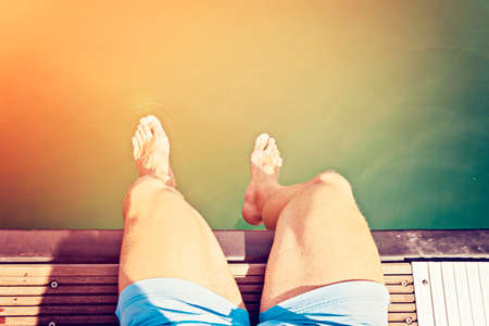 The man in blue shorts sits on the edge of the pier and holding his feet in the water, soft focus, light toned, defocused top viewの写真素材