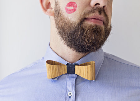 Close up Male cheek with red of female lips Bearded. Man in Blue Shirt and wooden Bow Tie with Red Lipstick on Cheek Happy Boss's Day Conceptの写真素材