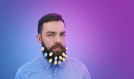 Portrait of handsome bearded man looking at camera with Easter eggs decoration in his beard on...
