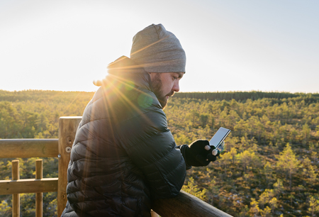 Bearded man looking at smartphone against autumn forest in sun beam outdoor lifestyle and smartphone conceptの写真素材