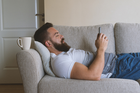 Close up portrait of adult caucasian bearded man lying on sofa and using smartphone. Internet addiction conceptの写真素材