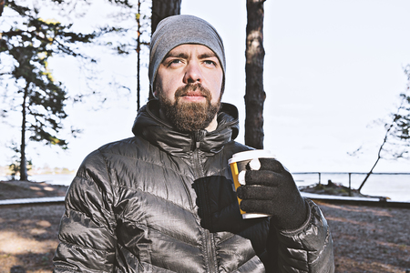 Caucasian bearded man with take away cup of coffee walking on sea coast and enjoys privacy and weather. Retreat and anti-stress conceptの写真素材