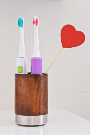 Two electric toothbrushes in wooden glass on sink in the bathroom with red heart. Personal hygiene like concept. Greeting card to celebrate Valentines dayの写真素材