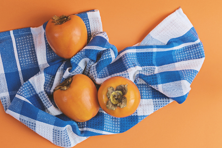 Three ripe persimmons on checkered blue towel on an orange background. Concept of autumn seasonal fruits, flat lay top viewの写真素材