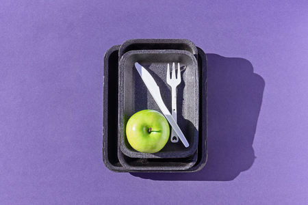 Top view of an empty plastic tray with green apple and disposable tableware on purple, flat lay. Concept of reducing the use of plasticの写真素材