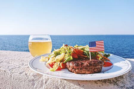 Plate with grilled juiciest burger meat and vegetables decorated with USA flag and glass of beer, closeup view, outdoor partyの写真素材