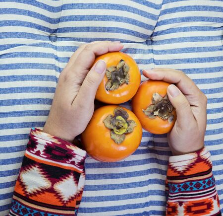 Female Hand holds three ripe persimmon on striped blue background. Minimal concept for decoration with copy spaceの写真素材