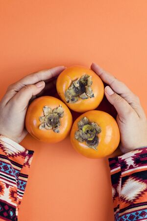Female Hand holds three ripe persimmon on orange background. Minimal concept for decoration with copy spaceの写真素材