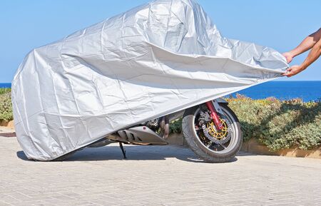 Biker covers motorcycle with waterproof cover with silver reflective surface protective. Motorbike covered with fabric shield parked at outdoorの写真素材