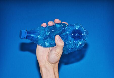 Hand squeezing plastic bottle on blue background. Zero waste and consumption reduction of plastic concept. Minimal flash photography, direct flash, copy spaceの写真素材