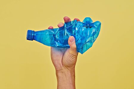 Hand squeezing blue plastic bottle on yellow background. Zero waste and consumption reduction of plastic concept. minimal, copy spaceの写真素材
