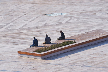 Three men are sitting on bench in public square observing social distance during pandemic of...
