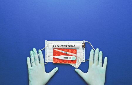 Hands in protective glove holds thermometer, syringe, facemask, pills and flu vaccine on blue.