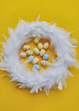 White feather wreath on yellow background with mini artificial eggs, top view. Easter decoration...