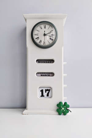 Wooden white vintage clock with calendar and Saint Patricks day cloverleaf on grey background