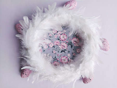 White feathers wreath on lilac floral background with decorative easter eggs, top view