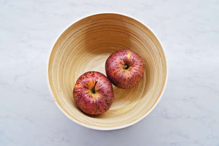Two red ripe apples in wooden bowl on a light background, top view