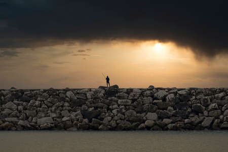 Lonely silhouette of fisherman with fishing rod against stormy sky on seashore at sunset, rear view