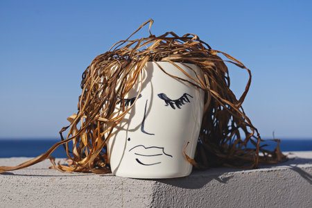 Ceramic pot with hand-drawn woman face and dried plant leaves mimicking woven hair sits against blue...