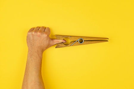 Creative composition of human hand gripped by large wooden clothespin, placed against vibrant yellow background, perfect for quirky visual conceptsの写真素材