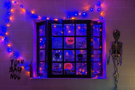 Halloween-themed window with spooky skeleton on glowing window, spooky lights in orange and blue illuminate creepy faces spooky vibe fills corner with festive mystery and night atmosphereの写真素材
