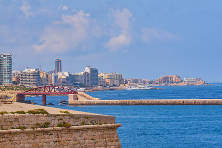 Coastal view of Valletta Malta with red breakwater bridge leading to harbor entrance and modern skyline under clear summer skyの写真素材