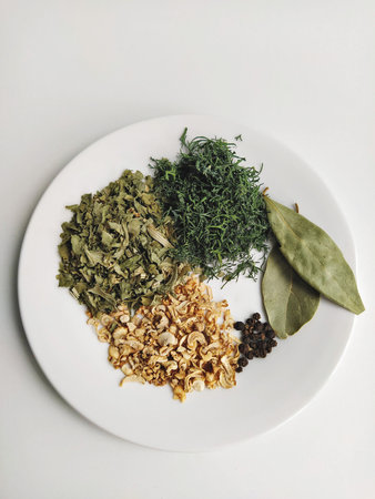 Assorted dried herbs on white plate: parsley, dill, celery, bay leaf and black pepper, clean overhead kitchen arrangementの写真素材