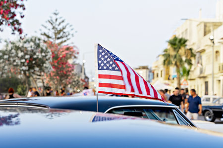 Small American flag on car roof at city street event, people and palm trees in soft background, bright summer vibeの写真素材