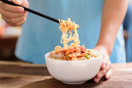 Korean food,hand holding chopsticks for eating instant noodle with kimchi cabbage in a bowl on wooden backgroundの写真素材