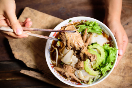 Rice noodles soup with stewed chicken wing and feet in a bowl and hand holding chopsticks for eatingの写真素材