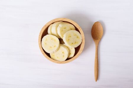 Sliced banana in a bowl and wooden spoon on white background, top viewの写真素材