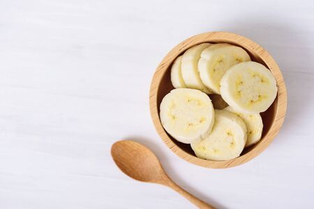 Sliced banana in a bowl and wooden spoon on white background, top viewの写真素材