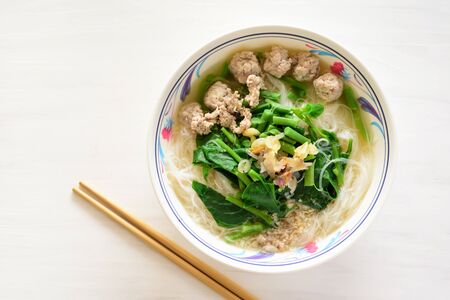 Rice noodles soup with pork meatball and vegetables in a bowl and chopsticks on white table, homemade Asian food, top viewの写真素材