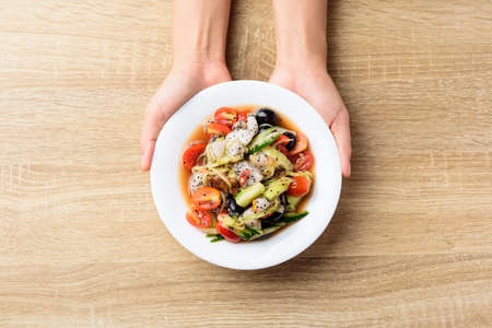 Spicy fruit and vegetables salad on white plate holding by woman hand on wooden table, Vegan foodの写真素材