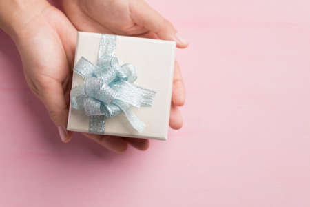 Silver gift box with light blue ribbon holding by hand on pink background, Present for giving in...