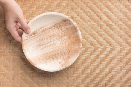 Hand holding Betel palm leaf plate (Biodegradable, Compostable or Eco friendly disposable plate) on woven bamboo sheet, sustainable conceptの写真素材