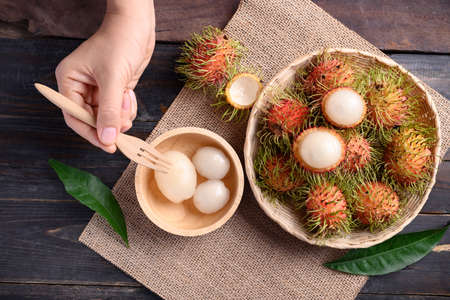 Woman hand holding fork for eating ripe rambutan fruit in a basket and bowl on wooden background, Rambutan is tropical fruit and native Southeast Asia, juicy and sweet tasteの写真素材