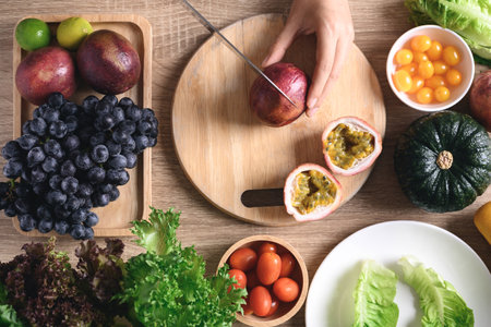 Hands of woman holding knife and cutting passion fruit on wooden board with fresh vegetables for vegan food cooking, Healthy eating, Table top viewの写真素材