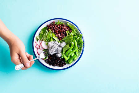 Healthy vegan salad vegetables with lettuce, corn salad, radish, kidney bean, azuki bean and green bean in a bowl on color background with hand holding fork for eatingの写真素材