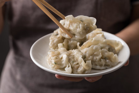Hand holding chopsticks and eating steamed dumpling filled with pork and vegetables on dishの写真素材