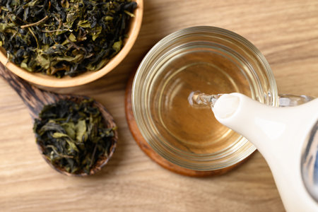 Hot green tea in teapot pouring into cup glass and dried tea leaves in bowl with spoon on wooden background, Healthy drink, Table top viewの写真素材