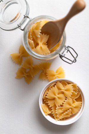 Top view of raw farfalle pasta in a white bowl and glass jar on white background, Bow-tie shaped pasta highlighting Italian culinary ingredientsの写真素材