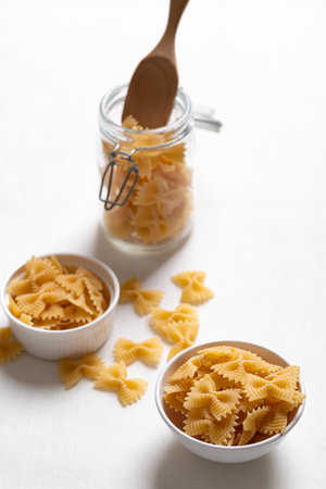 Raw farfalle pasta in a white bowl and glass jar on white background, Bow-tie shaped pasta highlighting Italian culinary ingredientsの写真素材