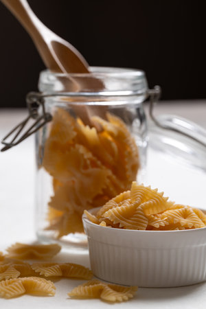 Raw farfalle pasta in a white bowl and glass jar with wooden spoon, Bow-tie shaped pasta highlighting Italian culinary ingredientsの写真素材