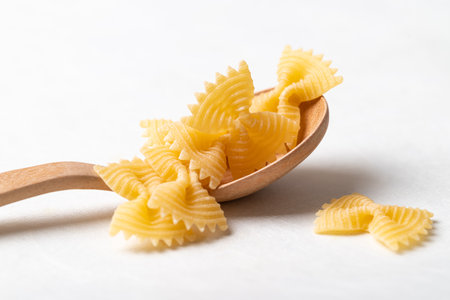 Raw farfalle pasta on a wooden spoon placed on a white background, Bow-tie shaped pasta highlighting Italian culinary ingredients and minimal food stylingの写真素材