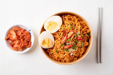 Spicy Korean dried instant noodles with boiled egg, sliced scallions, sesame seed and fresh chili in kraft paper bowl on white backgroundの写真素材