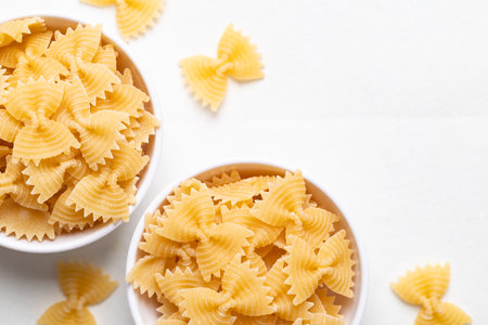 Top view of raw farfalle pasta in a simple white bowl on a clean white background. Bow-tie shaped pasta highlighting Italian culinary ingredientsの写真素材