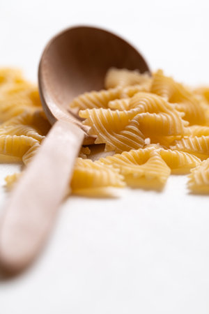 Raw farfalle pasta with wooden spoon on white background, Bow-tie shaped pasta highlighting Italian culinary ingredientsの写真素材