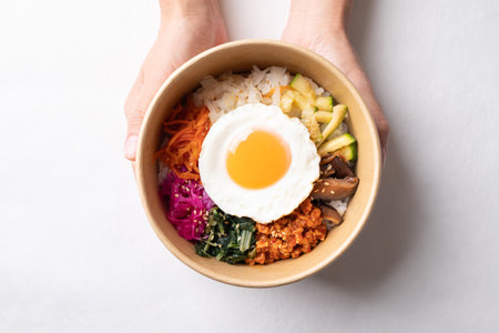 Bibimbap, Korean food, Cooked rice with vegetables, pork and egg in biodegradable bowl holding by hand on white background, Top viewの写真素材